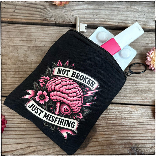 A black canvas zippered pouch with a pink and white graphic of a brain, flowers, and the text 'NOT BROKEN JUST MISFIRING', shown on a rustic wooden surface with a key and small items peeking out from the top.