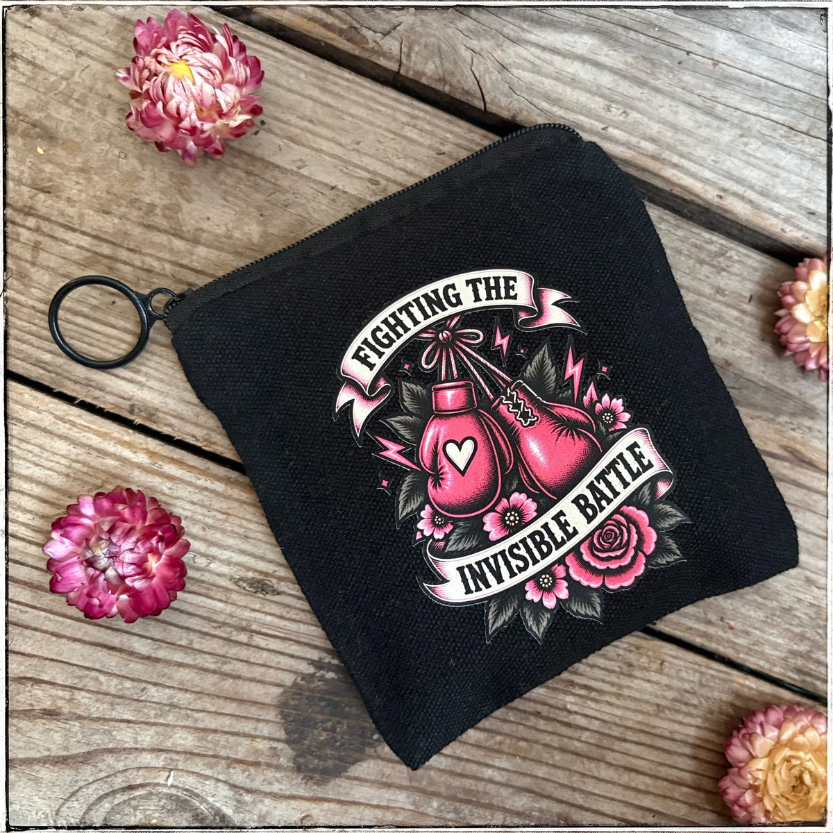 A black canvas zip pouch with a graphic print of pink boxing gloves, roses, and banners reading 'FIGHTING THE INVISIBLE BATTLE,' photographed on a rustic wooden surface with pink flowers around it.