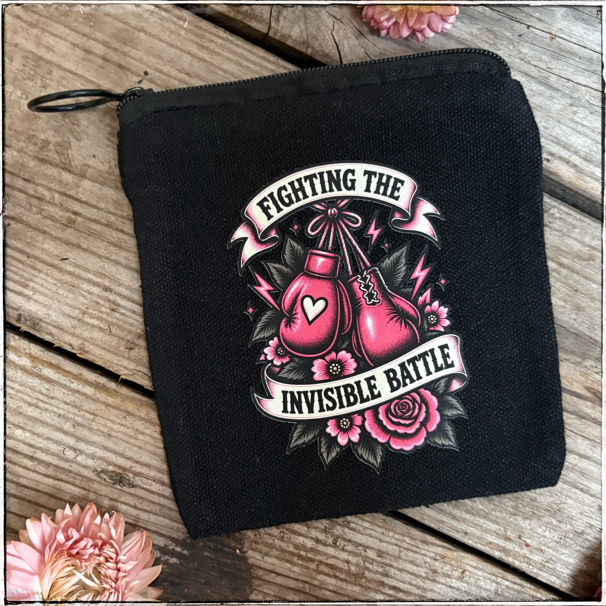 A black canvas zippered pouch featuring a graphic print of two pink boxing gloves, roses, and banners that read 'FIGHTING THE INVISIBLE BATTLE.' The pouch is photographed on a rustic wooden surface with pink flowers placed around it.
