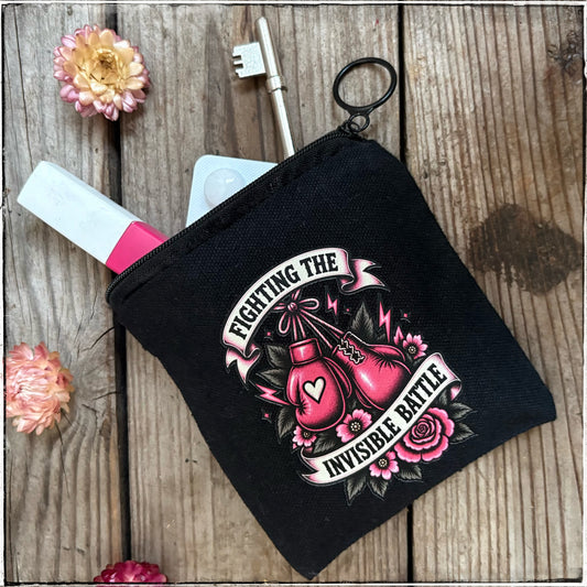 A black canvas coin pouch with a zipper closure, featuring a graphic print of pink boxing gloves and roses with the text 'FIGHTING THE INVISIBLE BATTLE'. The pouch is styled on a rustic wooden background with decorative flowers and a key.