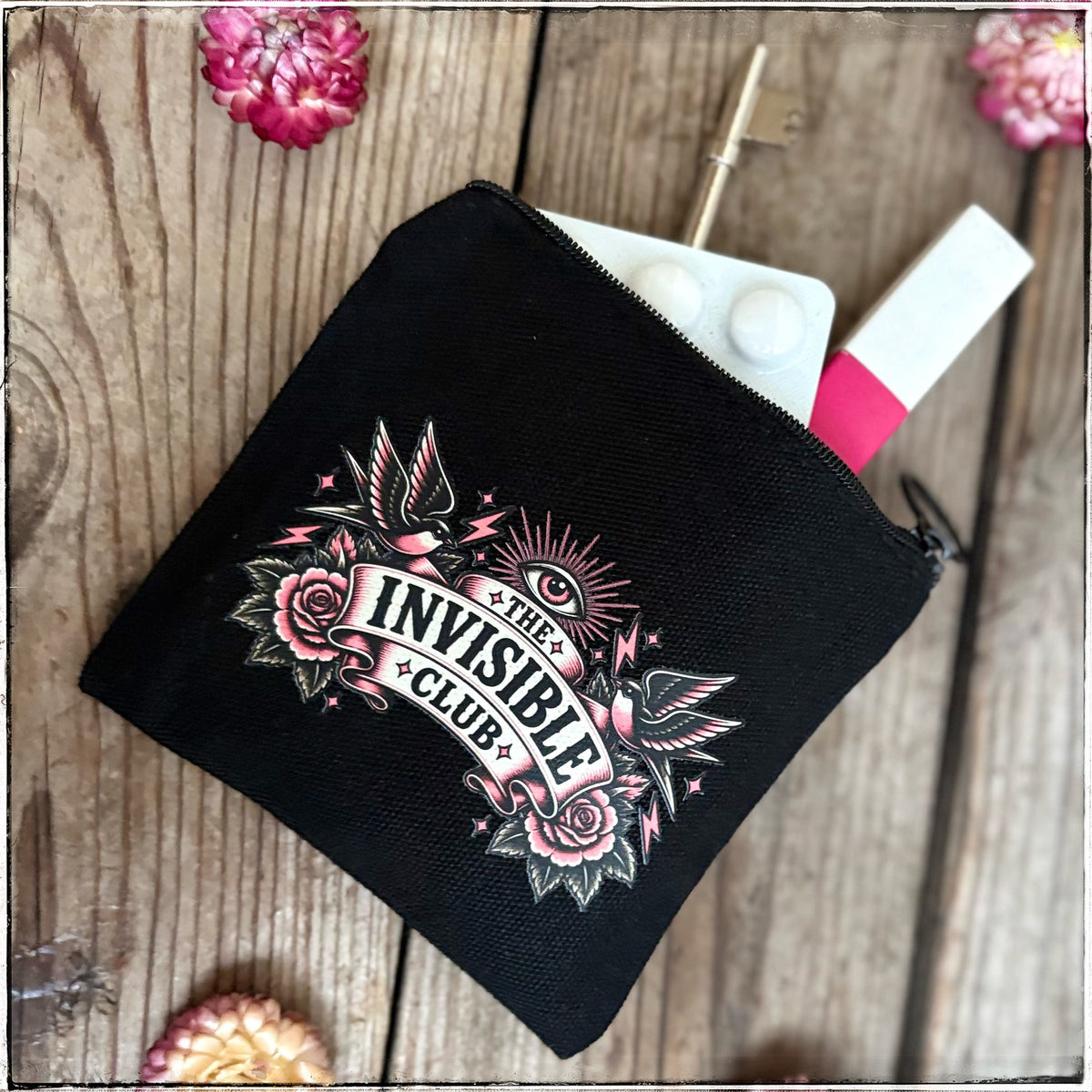 A black canvas zippered pouch with a tattoo-style graphic reading 'THE INVISIBLE CLUB' in pink, white, and black, shown on a rustic wooden surface with dried flowers and small accessories peeking out from inside.