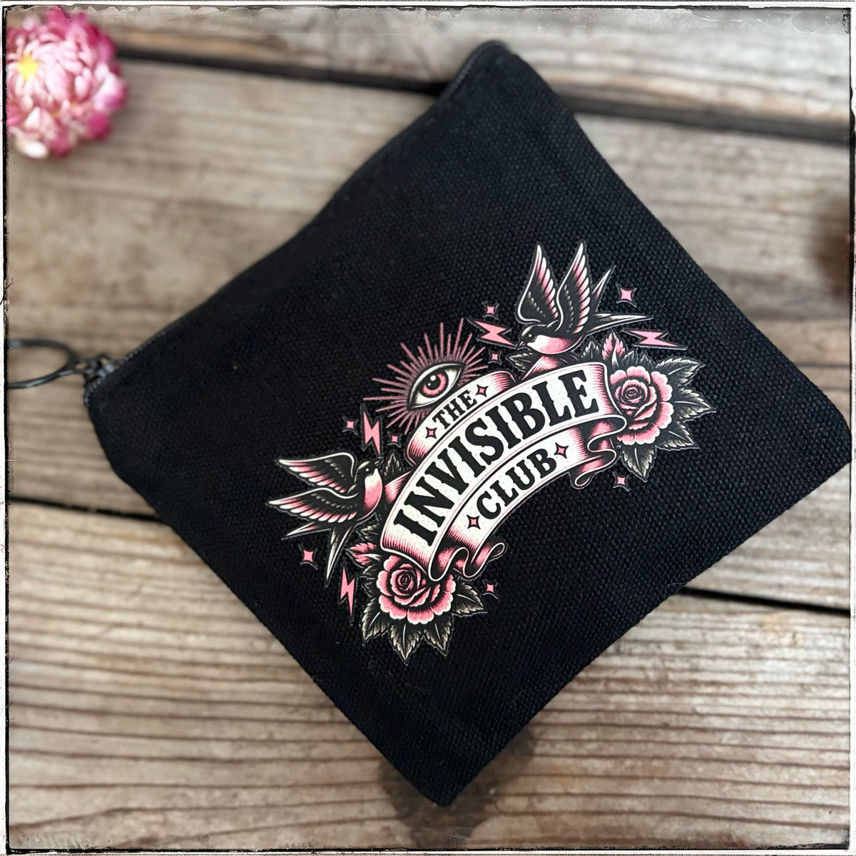 A black canvas zippered pouch featuring a tattoo-style graphic that reads 'THE INVISIBLE CLUB' with pink roses, birds, and an eye motif, photographed on a rustic wooden surface.