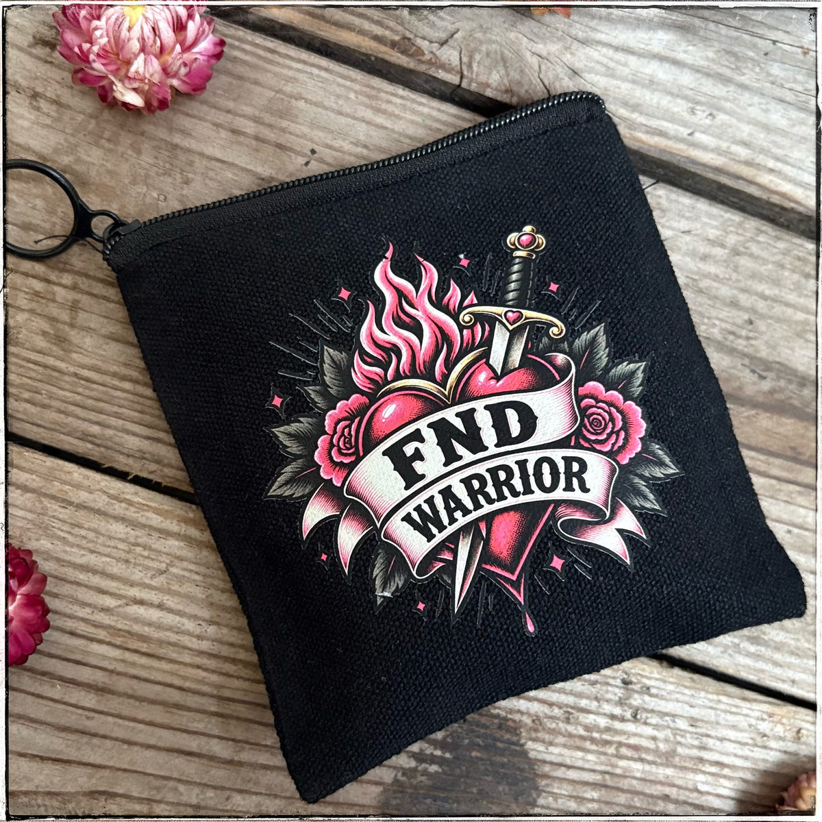 A black canvas zippered pouch with a key ring, featuring a traditional tattoo-style graphic of a heart pierced by a sword with flames and roses, and a banner reading 'FND WARRIOR'. The pouch is displayed on a rustic wooden surface with pink flowers in the corners.