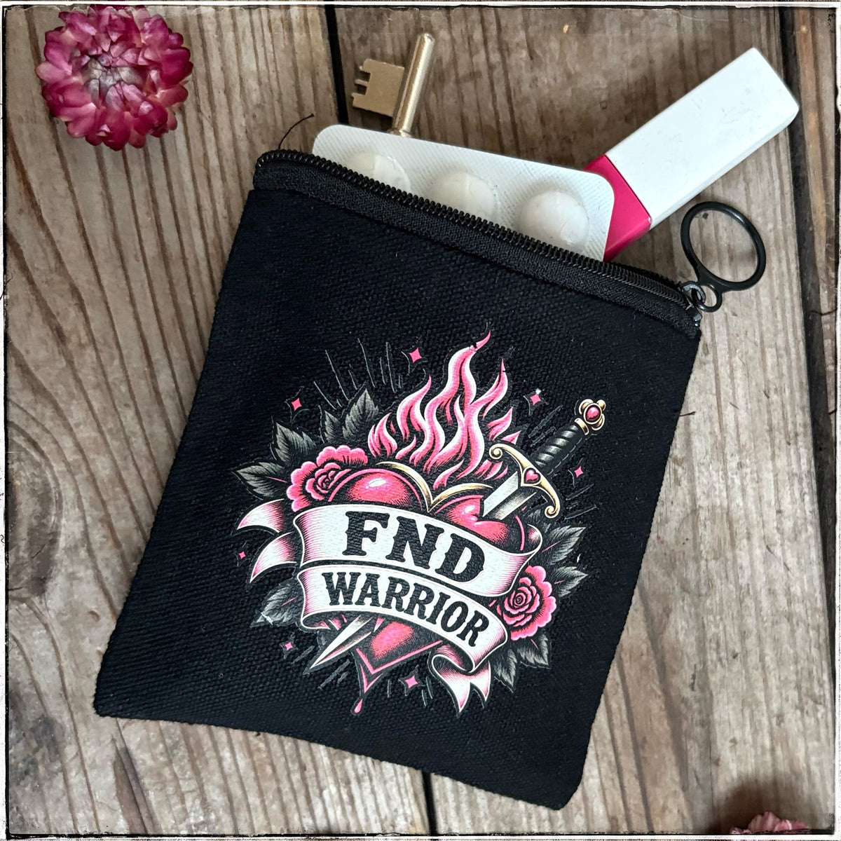 A black canvas zippered pouch with a colorful tattoo-style graphic reading 'FND WARRIOR' on the front, shown on a rustic wooden surface with a key, pill bottle, and small flower prop.