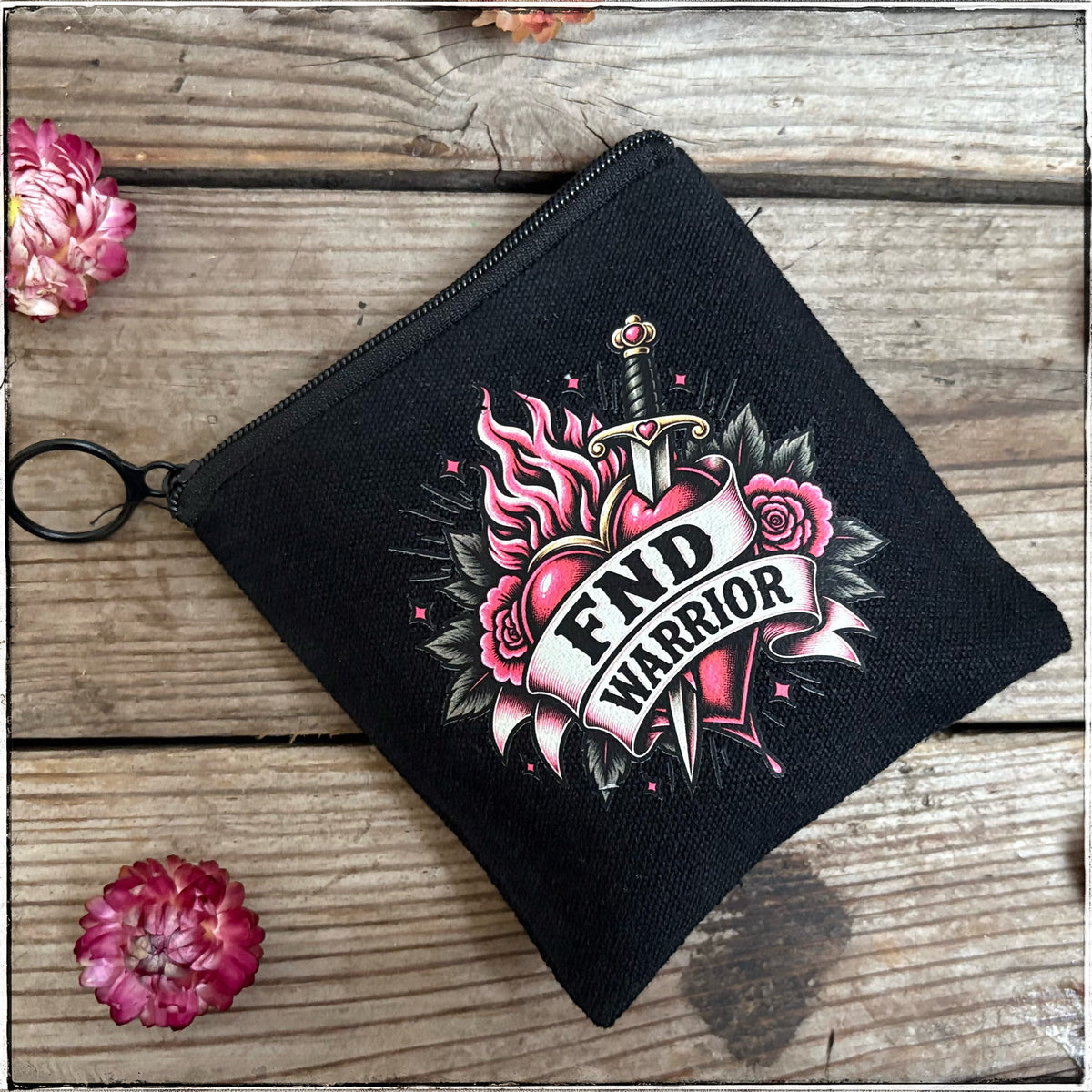 A black canvas zippered pouch with a graphic print featuring a dagger piercing a heart, pink roses, and the words 'FIND WARRIOR' in a banner. The pouch is displayed on a rustic wooden surface with two pink flowers in the corners.