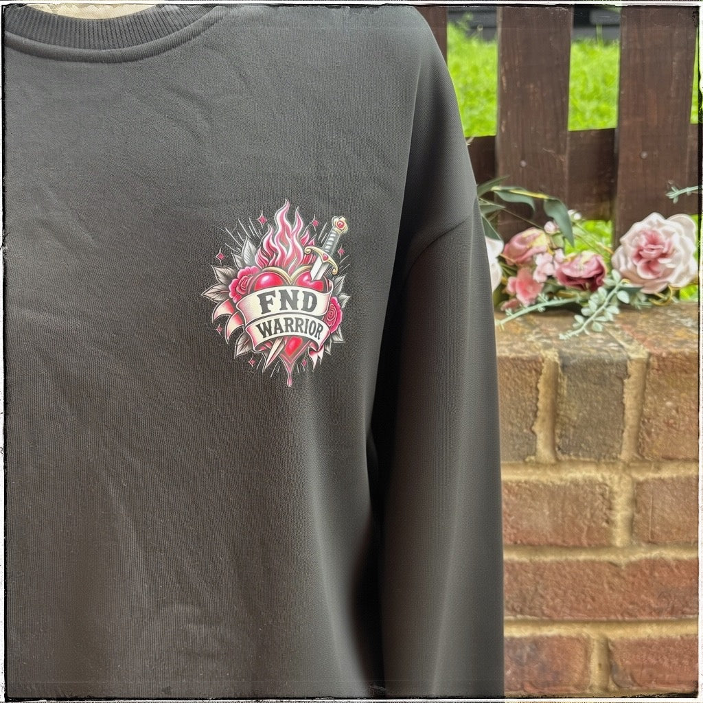 A black sweatshirt featuring a pink and white 'FND WARRIOR' graphic on the left chest, and displayed against an outdoor brick wall background with flowers.