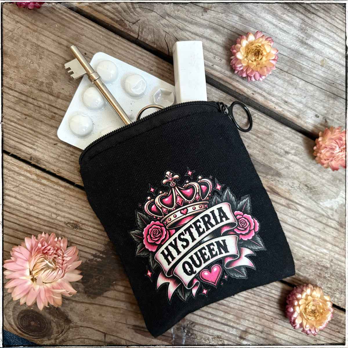 A black canvas zippered pouch with a 'Hysteria Queen' graphic print, styled on a rustic wooden background with pink flowers and small props peeking out from the top.
