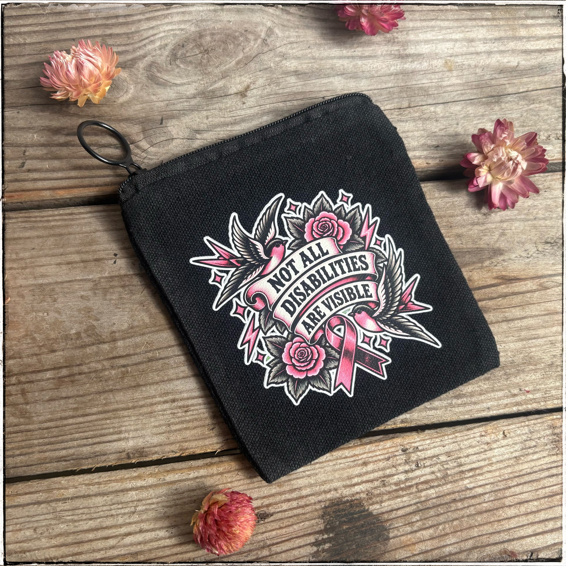 A black canvas zippered pouch with a black metal key ring, featuring a pink and white graphic design with roses, birds, and the text 'Not All Disabilities Are Visible’. The pouch is displayed on a rustic wooden surface decorated with dried pink flowers.