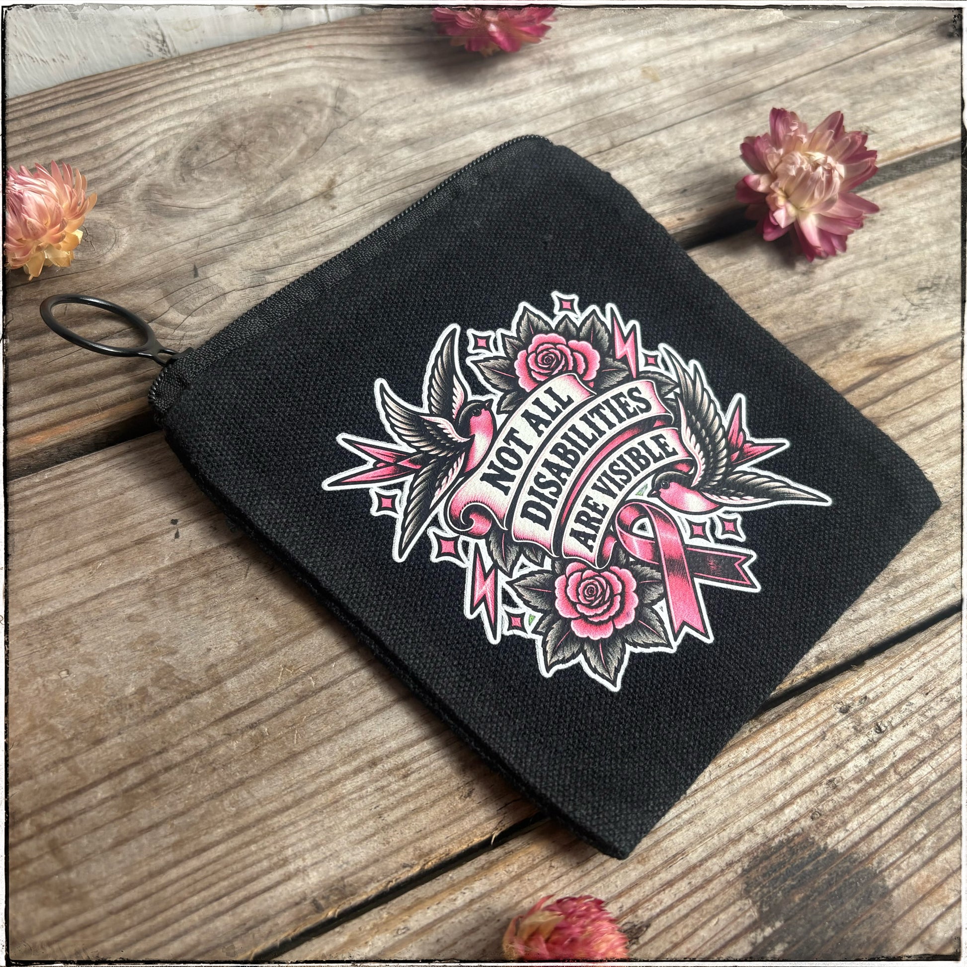 A black canvas zippered pouch with a pink and white graphic that reads 'Not All Disabilities Are Visible,' displayed on a rustic wooden surface with dried flowers.