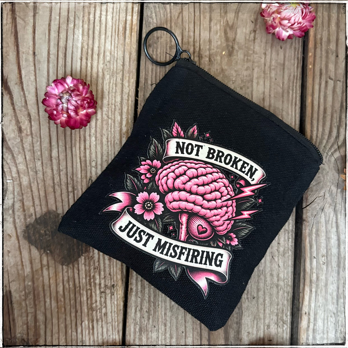A black canvas zippered pouch with a key ring, featuring a pink and white tattoo-style graphic of a brain with flowers and the text 'NOT BROKEN JUST MISFIRING', displayed on a rustic wooden background with decorative pink flowers.