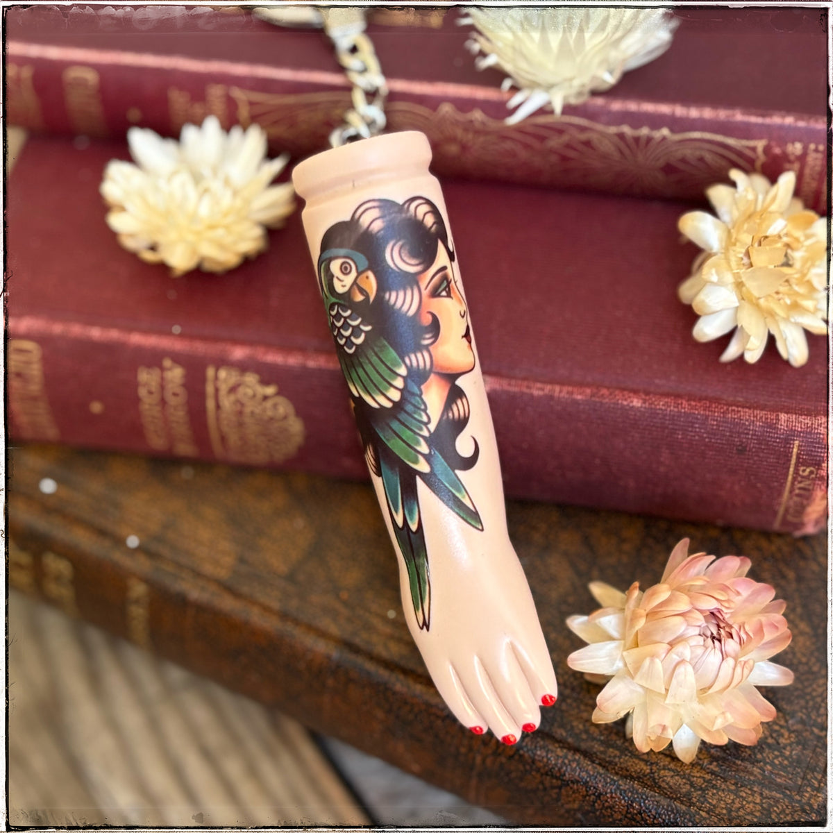 A porcelain doll arm with red toenails and a tattoo-style illustration of a woman's face and green parrot, shown resting on stacked vintage books with dried flowers.