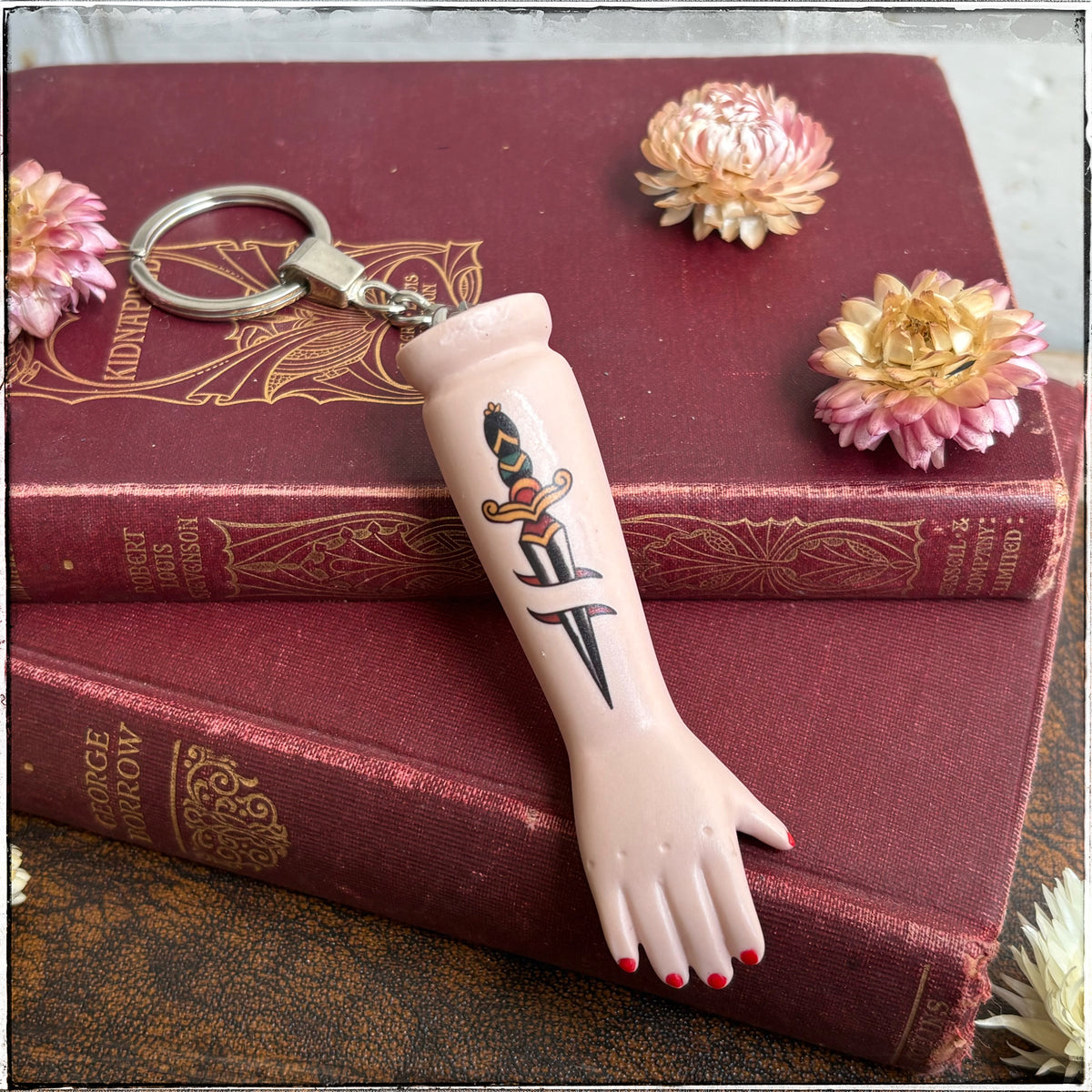A recycled porcelain doll arm keychain with a tattoo of a dagger and red fingernails, resting on stacked vintage red books with decorative flowers