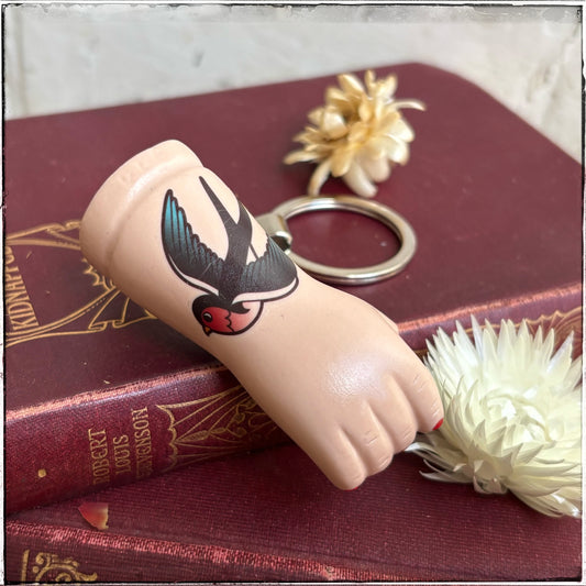 A recycled porcelain doll arm keychain with a tattoo of a swallow and red fingernails, resting on stacked vintage red books with decorative flowers