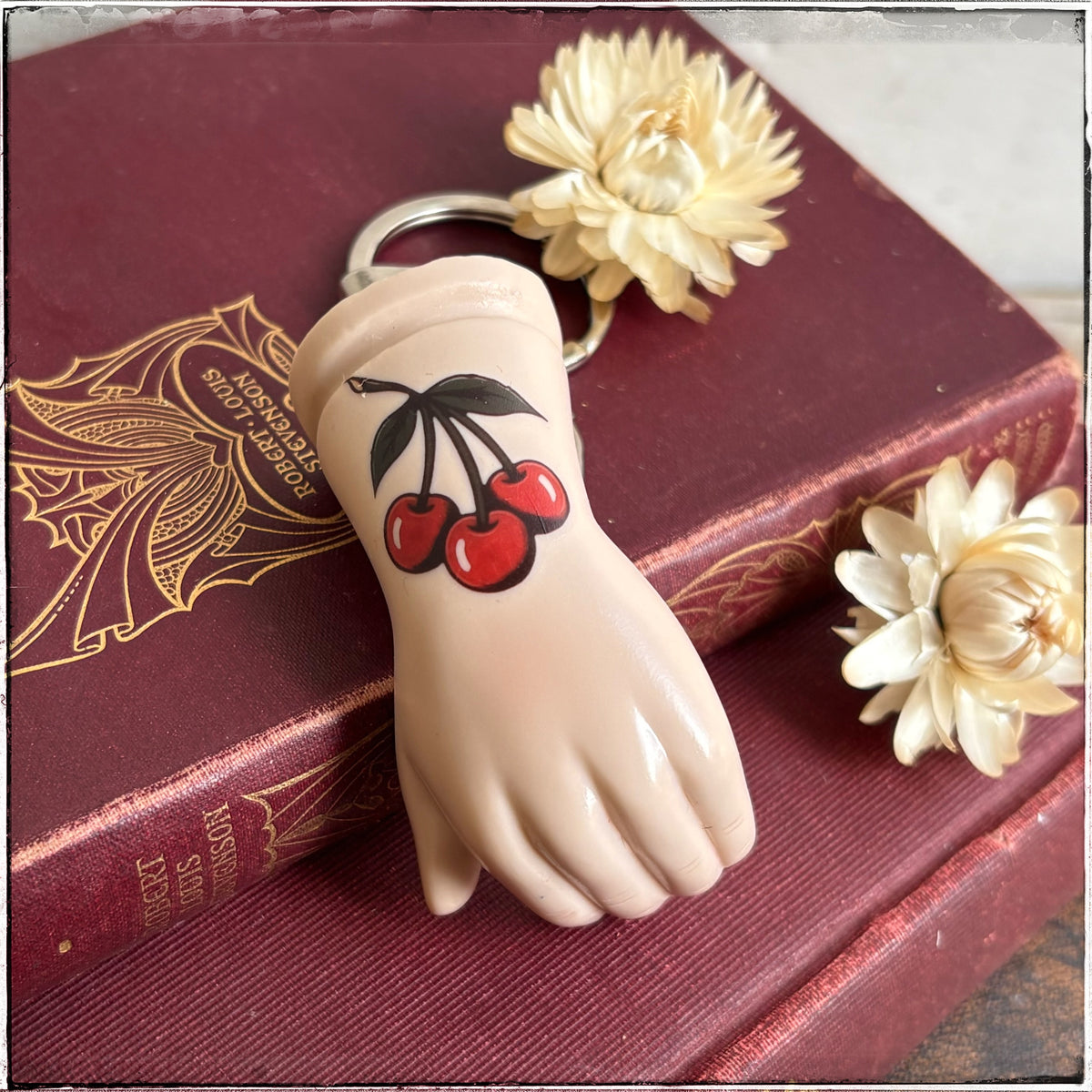 A porcelain hand-shaped keychain with a cherry design, resting on a vintage red book with cream flowers.