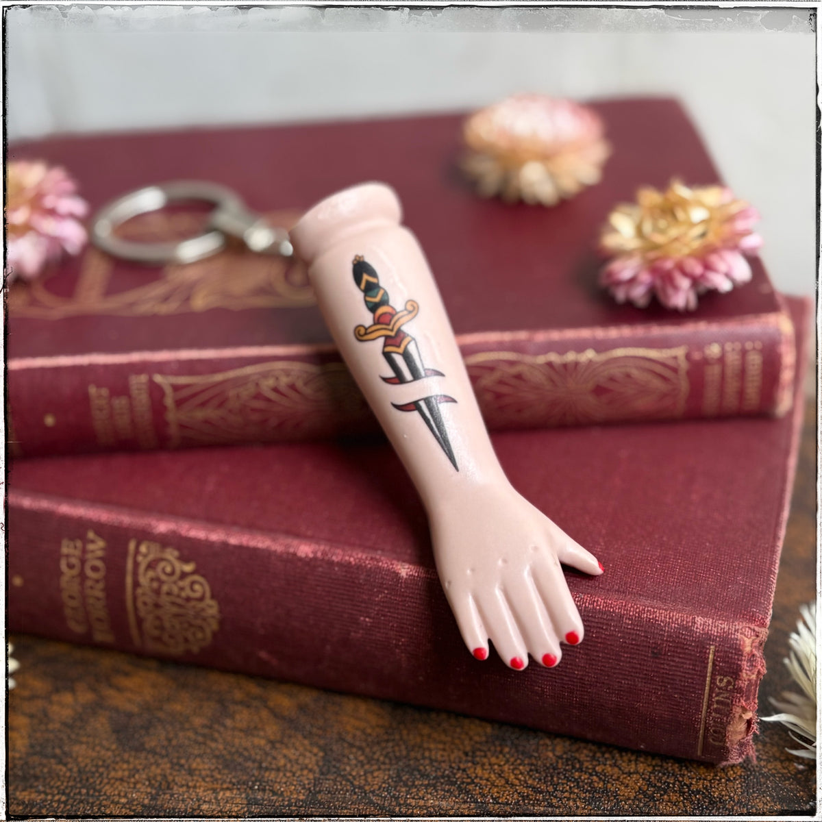 A recycled porcelain doll arm keychain with a tattoo of a dagger and red fingernails, resting on stacked vintage red books with decorative flowers.