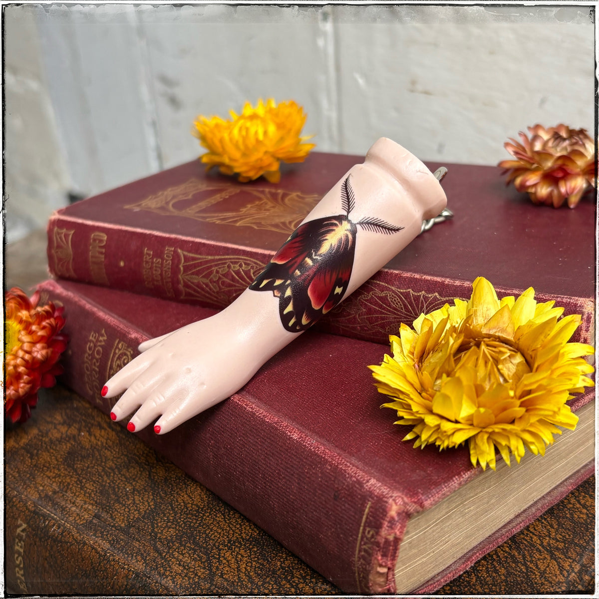 A recycled porcelain doll arm keychain with a tattoo of a moth tattoo and red fingernails, resting on stacked vintage red books with decorative flowers