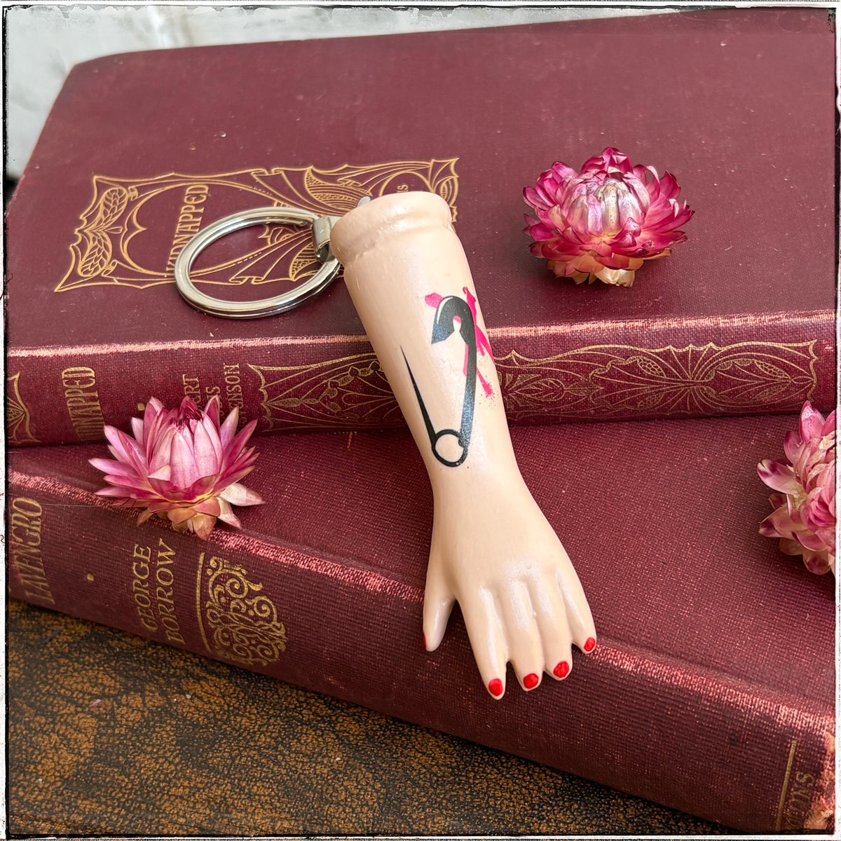 A recycled porcelain doll arm keychain with a tattoo of a safety pin and red fingernails, resting on stacked vintage red books with decorative flowers