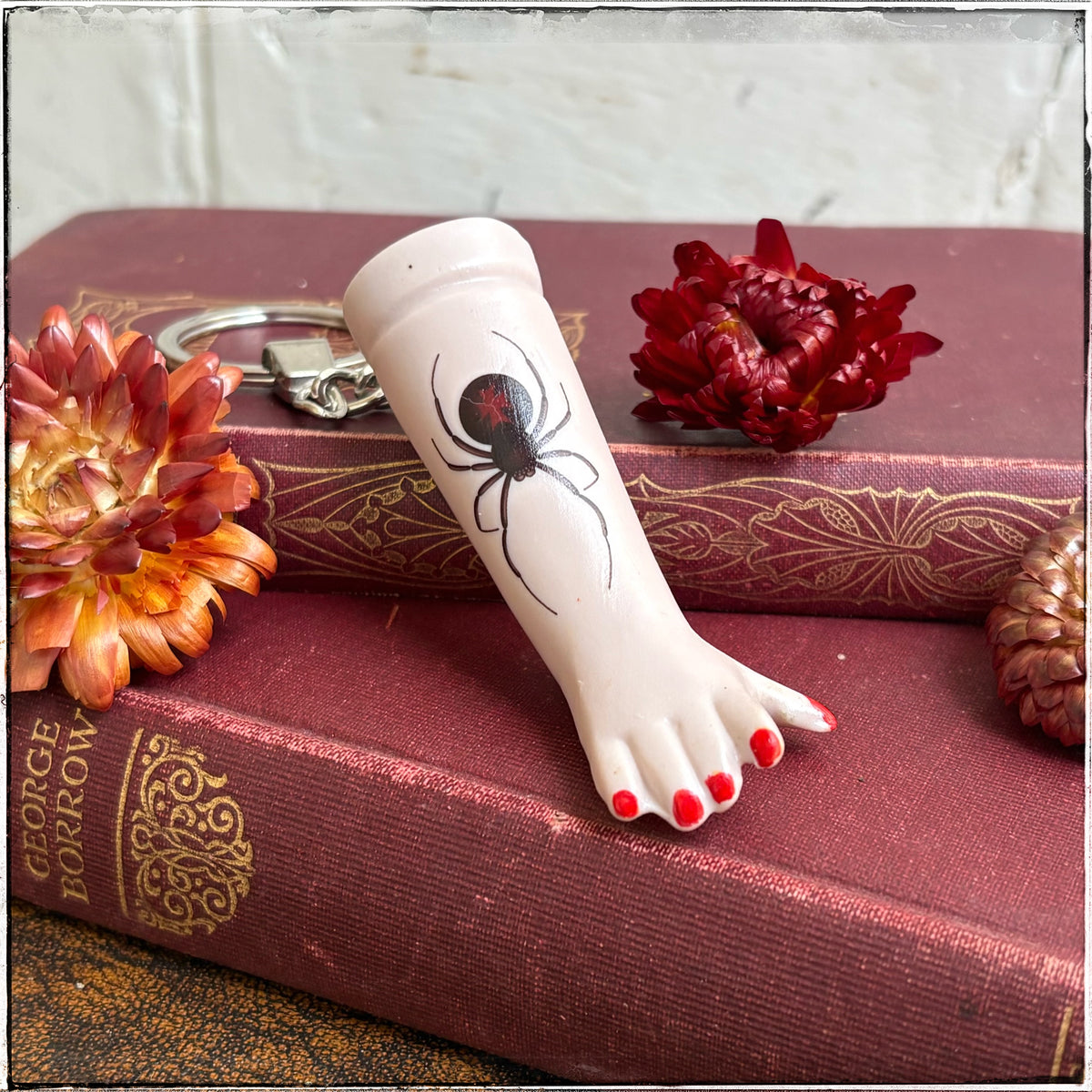 A recycled porcelain doll arm keychain with a tattoo of a spider and red fingernails, resting on stacked vintage red books with decorative flowers.
