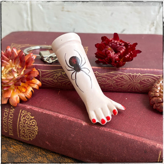 A recycled porcelain doll arm keychain with a tattoo of a spider and red fingernails, resting on stacked vintage red books with decorative flowers.
