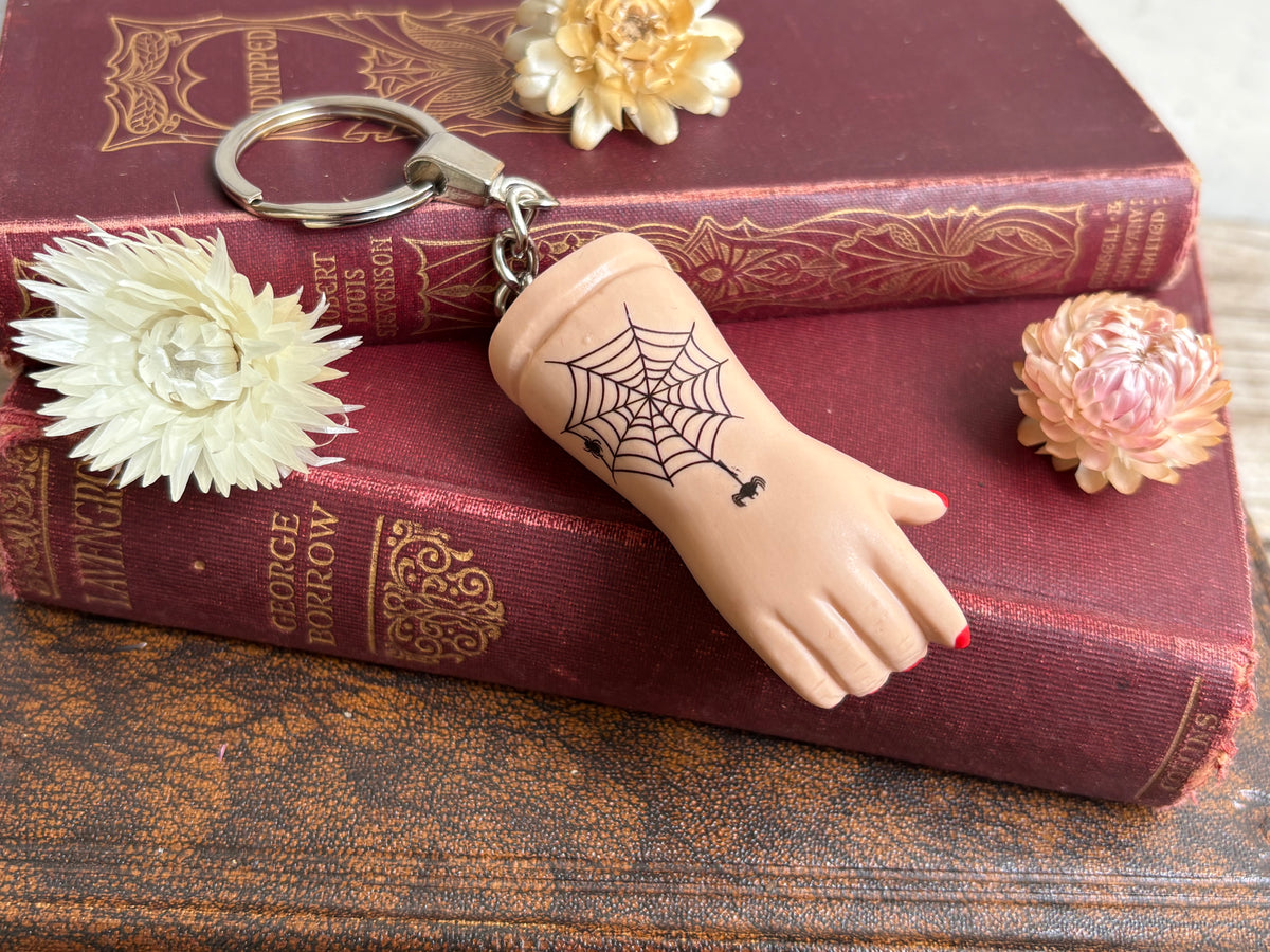 A recycled porcelain doll arm keychain with a tattoo of a spiderweb tattoo and red fingernails, resting on stacked vintage red books with decorative flowers