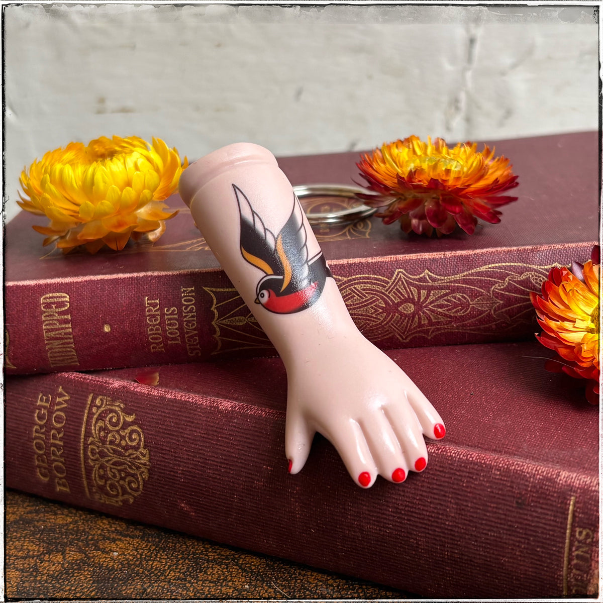 Swallow Tattoo Porcelain Doll Arm Keyring 

A recycled porcelain doll arm keychain with a tattoo of a swallow and red fingernails, resting on stacked vintage red books with decorative flowers
