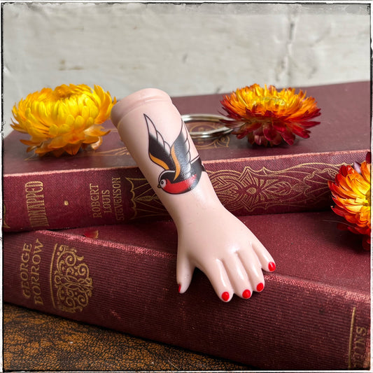 Swallow Tattoo Porcelain Doll Arm Keyring 

A recycled porcelain doll arm keychain with a tattoo of a swallow and red fingernails, resting on stacked vintage red books with decorative flowers
