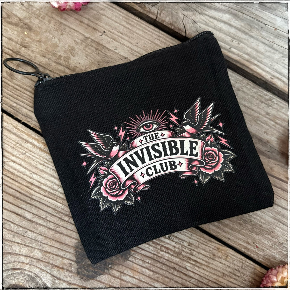 A black canvas zippered pouch featuring a gothic tattoo-style graphic that reads 'THE INVISIBLE CLUB' with pink roses, a bird, and an eye, placed on a rustic wooden surface.
