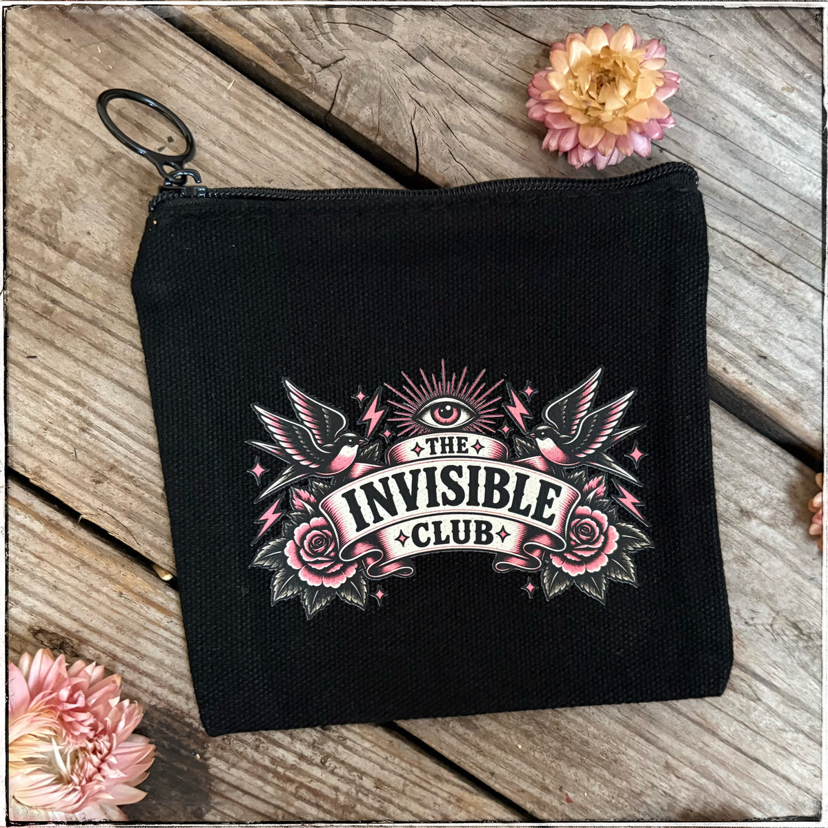 A black zippered pouch with a key ring, featuring a pink and white graphic that reads 'THE INVISIBLE CLUB' with roses, birds, and stars, displayed on a rustic wooden background with pink flowers.